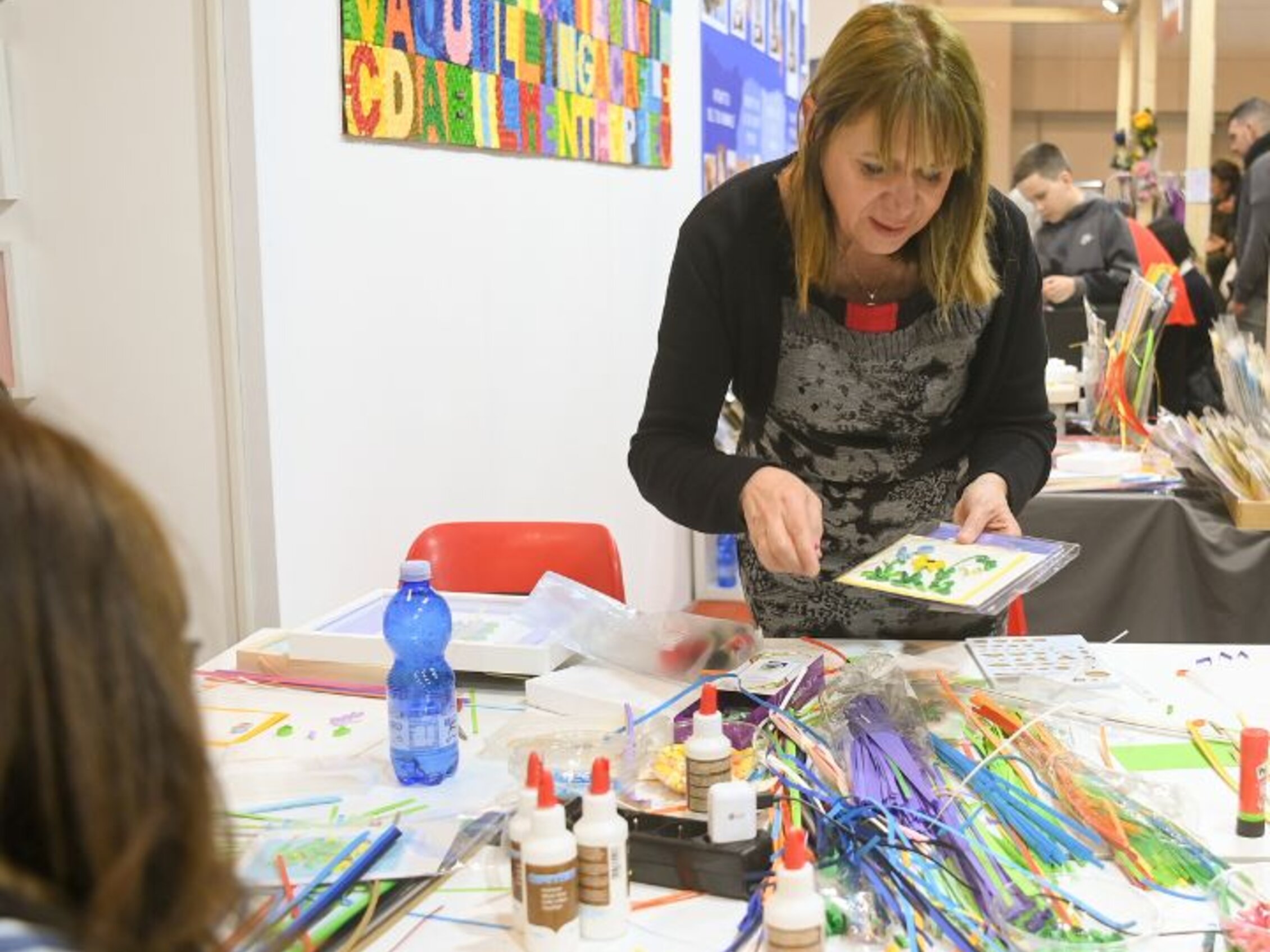 Insegnante mostra tecniche di quilling a un gruppo di partecipanti seduti attorno a un tavolo pieno di materiali creativi, nell’area Edu Lab della fiera Abilmente