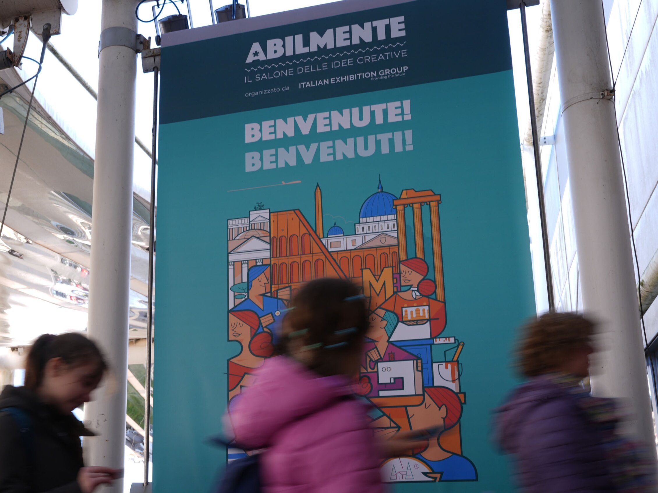 Ingresso di Abilmente Roma con cartellone di benvenuto e visitatori in arrivo alla fiera creativa.