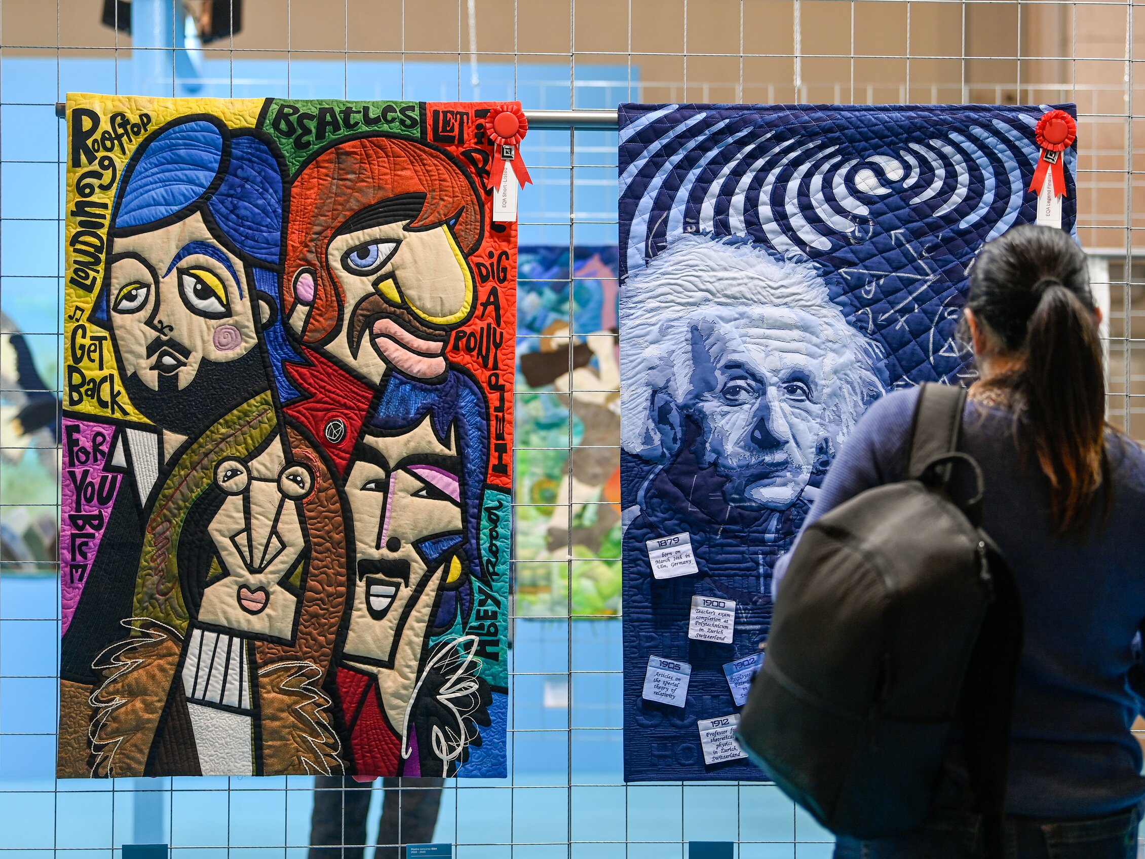 Visitatrice osserva due quilt artistici premiati: uno ritrae Einstein in toni blu con spirale ipnotica, l'altro i Beatles in stile pop cubista, esposti ad Abilmente. | © © Abilmente / Italian Exhibition Group – Foto di opere di quilt art premiate ed esposte nell’area creativa della fiera