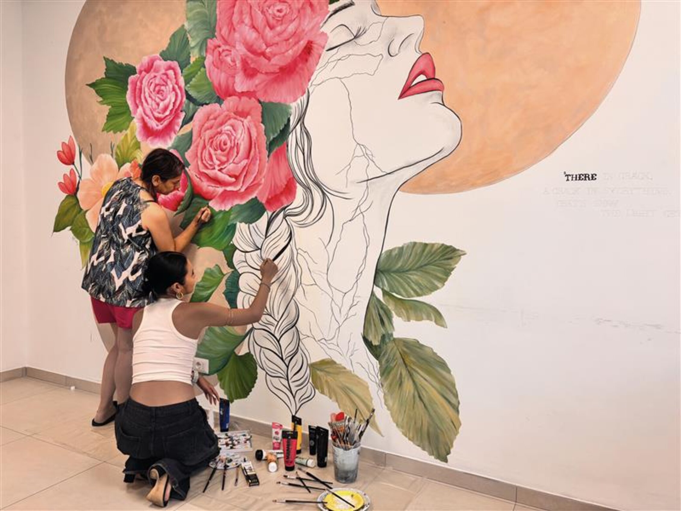 Due donne dipingono un grande murale su parete raffigurante un volto femminile con capelli intrecciati, labbra rosse e fiori colorati, parte della mostra inclusiva framMenti d’arte ad Abilmente Roma