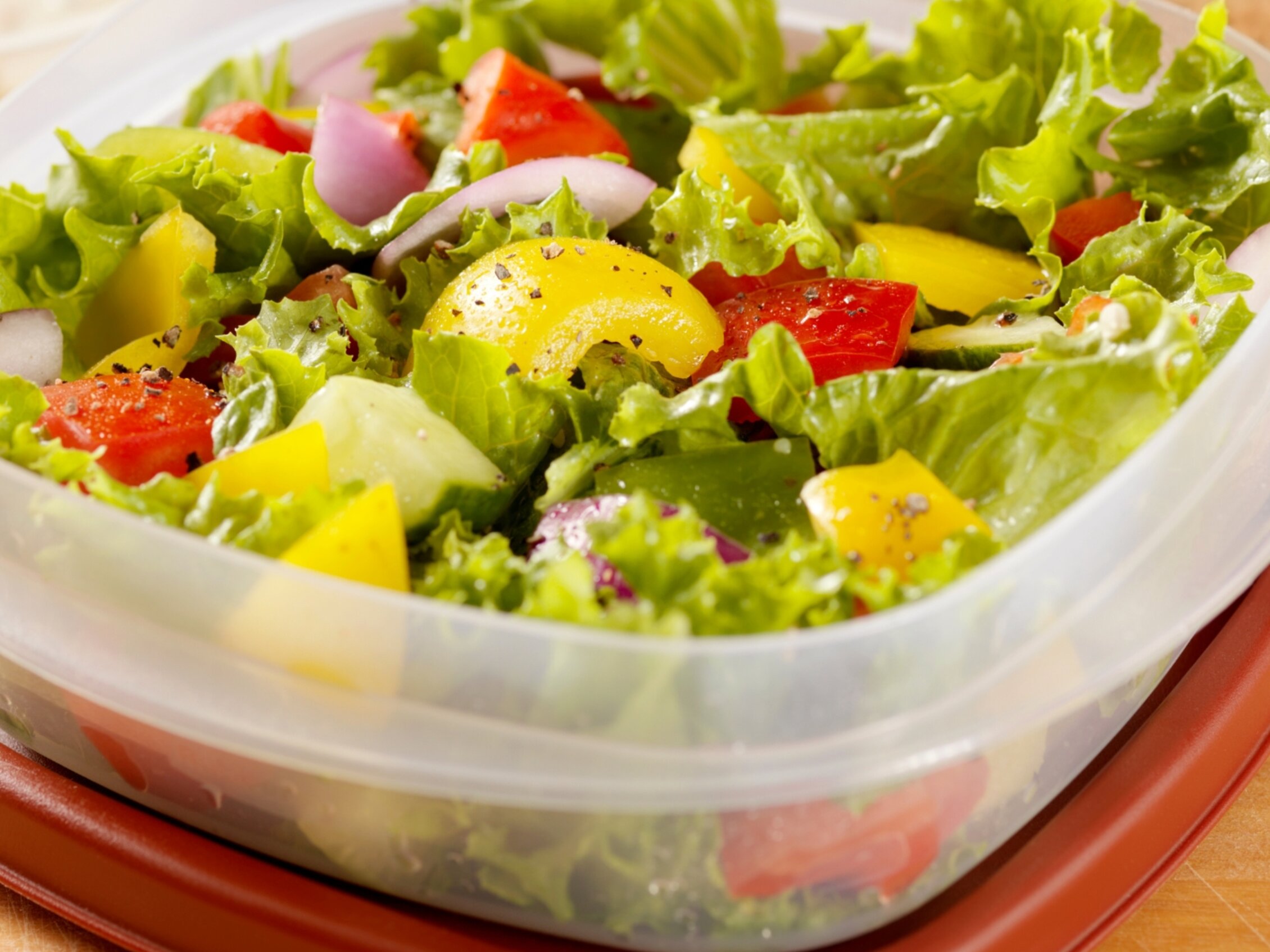 Insalata fresca colorata in contenitore riutilizzabile portata da casa per pic nic in fiera | © Primo piano di un’insalata mista fresca in contenitore di plastica trasparente con coperchio rosso: lattuga verde, pomodori, peperoni gialli e rossi, cipolla rossa e spezie sparse in superficie.