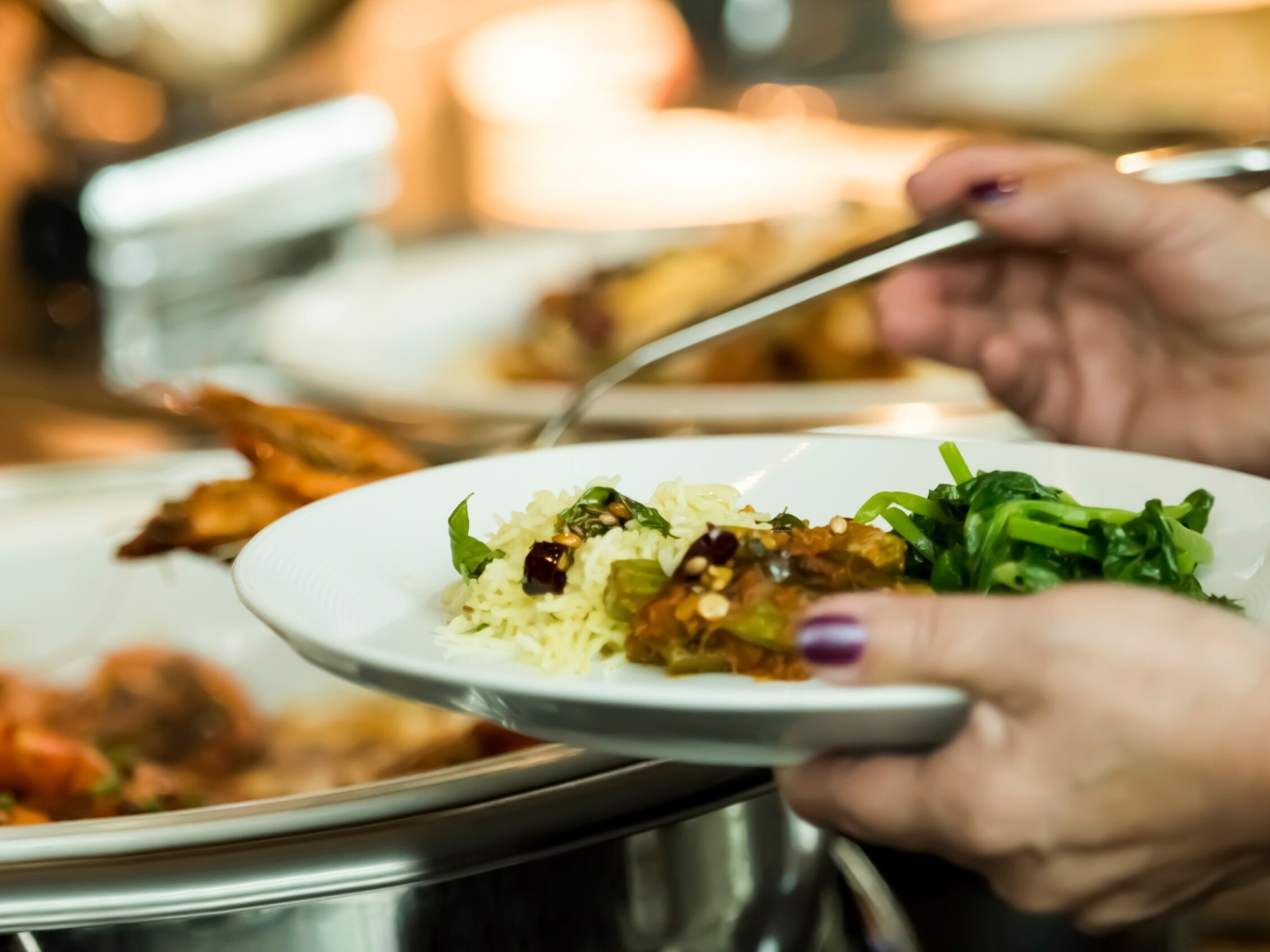 Primo piano di mano che impugna un piatto bianco con riso basmati, verdure al vapore e pietanza in umido, mentre si serve da un buffet con coperchi in acciaio lucido.